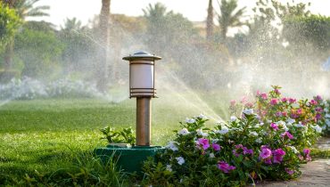 Plastic sprinkler irrigating flower bed on grass lawn with water in summer garden.